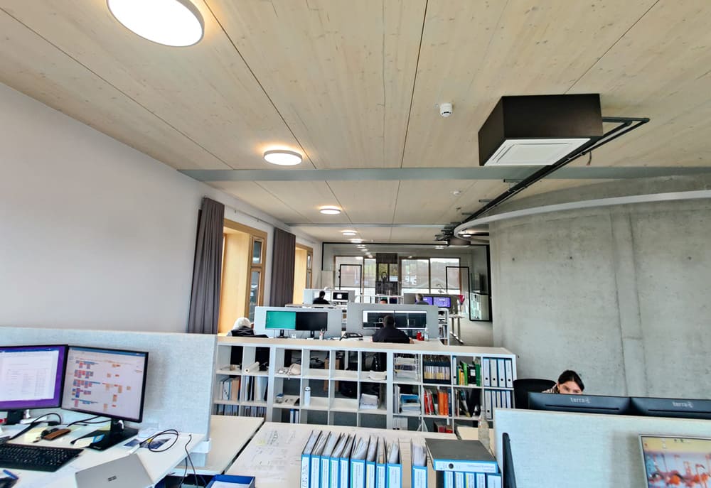 Büro mit mehreren Arbeitsplätzen, Regalen mit Ordnern und großen Monitoren, sichtbare Deckenbeleuchtung und Betonwand